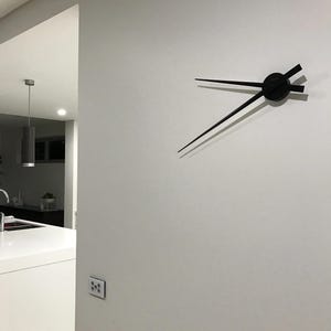 Peut inclure: Une horloge murale moderne et minimaliste avec des aiguilles noires et un centre circulaire. Les aiguilles sont longues et fines, avec un design épuré. L'horloge est fixée sur un mur uni de couleur claire, créant un contraste saisissant.