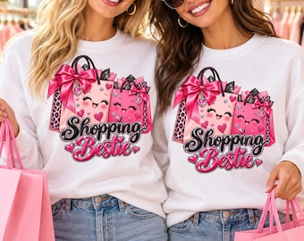 Könnte beinhalten: Ein weißes Langarm-Sweatshirt mit einem Grafikdruck von zwei rosa Einkaufstaschen mit lächelnden Gesichtern, großen rosa Schleifen und Leopardenmuster-Akzenten. Der Text Shopping Bestie ist in einer fetten, schwarzen und rosa Schrift gedruckt.