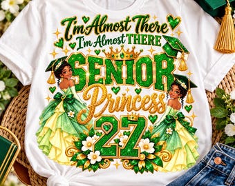 Könnte beinhalten: Ein weißes T-Shirt mit einem Grafikdruck und dem Text I'm Almost There Senior Princess 27. Das Design zeigt zwei Illustrationen einer Prinzessin in einem grün-gelben Kleid mit Doktorhut, umgeben von grünen Glitzerakzenten und Blumen.