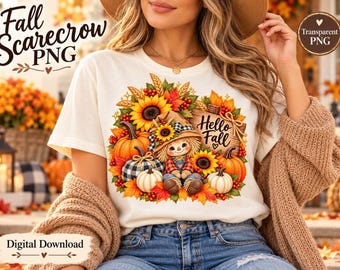 Könnte beinhalten: Ein cremefarbenes T-Shirt mit einem Grafikdruck einer Vogelscheuche, umgeben von Kürbissen, Sonnenblumen und Herbstblättern. Ein Holzschild rechts neben der Vogelscheuche zeigt den Text Hello Fall in schwarzer Schreibschrift.
