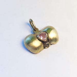 May include: A gold-toned pendant in the shape of a bow with a pink heart-shaped stone in the center. The pendant has a loop at the top for a chain. The bow has a textured surface.