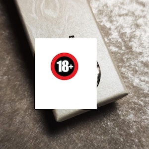Könnte beinhalten: Eine rechteckige, hellbeige strukturierte Schachtel mit Klappdeckel. Ein weißer quadratischer Aufkleber befindet sich in der Mitte der Schachtel und zeigt einen roten kreisförmigen Rand um den schwarzen Text 18+ in einer serifenlosen Schrift.