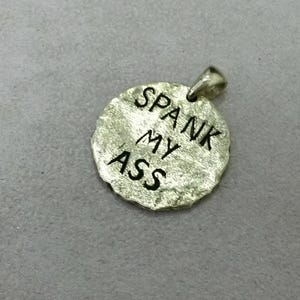 Könnte beinhalten: Ein silberfarbener Anhänger mit den Worten "SPANK MY ASS" in schwarzer, stilisierter Schrift graviert. Der Anhänger hat eine strukturierte, unregelmäßige Oberfläche und eine kleine Schlaufe für eine Kette.