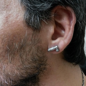 May include: A silver-colored earring in the shape of a handgun is worn in the ear. The earring is detailed and appears to be made of metal. The image shows a close-up of the ear and part of the face.