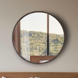 May include: A round mirror with a dark brown frame hangs above a bed with a light brown headboard. The bed has white bedding and a light brown blanket. Two nightstands flank the bed, and a vase of dried plants sits on one.