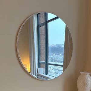 Może przedstawiać: Okrągłe lustro w srebrnej ramie wisi na beżowej ścianie. Poniżej biała komoda z sześcioma szufladami i kryształowymi gałkami. Na komodzie stoi biały wazon. Lustro odbija okno.