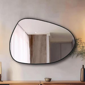May include: A modern living room featuring a large, irregularly shaped mirror with a black frame. The room includes a wooden console table, two pendant lights with spherical shades, and decorative vases. The color palette is neutral with warm wood tones.