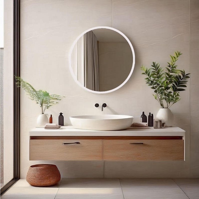A modern bathroom vanity features a round white mirror and a light wood floating cabinet with two drawers. A white oval sink sits atop the cabinet.