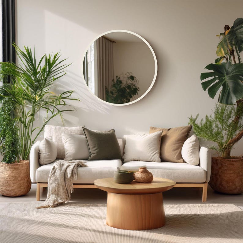 A living room scene featuring a cream-colored sofa with several pillows in neutral tones. A round wooden coffee table sits in front of the sofa, and a large round mirror hangs on the wall. Potted plants add a touch of greenery.