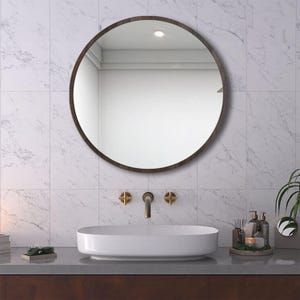 A round, dark-framed mirror hangs above a white oval sink in a bathroom setting. The mirror reflects the room's interior. The sink is paired with gold-toned faucets and is set against a marble-patterned tile wall.