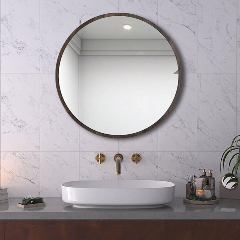 A round, dark-framed mirror hangs above a white oval sink in a bathroom setting. The mirror reflects the room's interior. The sink is paired with gold-toned faucets and is set against a marble-patterned tile wall.