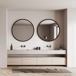 A modern bathroom design featuring two round mirrors with black frames above a double vanity. The vanity has a light-colored countertop and wooden cabinets. The room has a neutral color palette with a wooden floor and a dark gray bath mat.