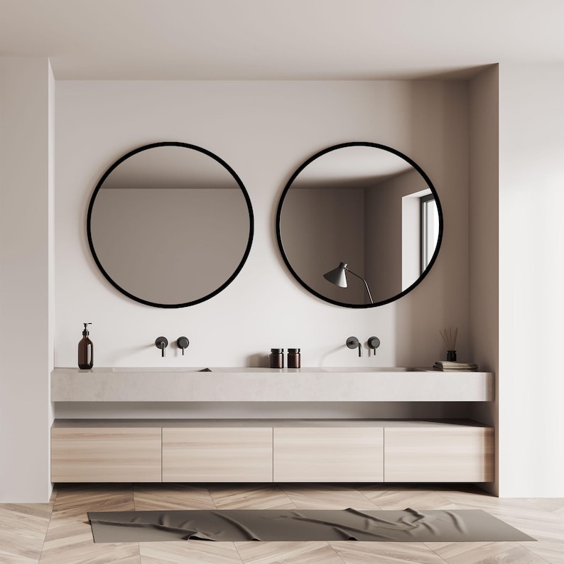 A modern bathroom design featuring two round mirrors with black frames above a double vanity. The vanity has a light-colored countertop and wooden cabinets. The room has a neutral color palette with a wooden floor and a dark gray bath mat.