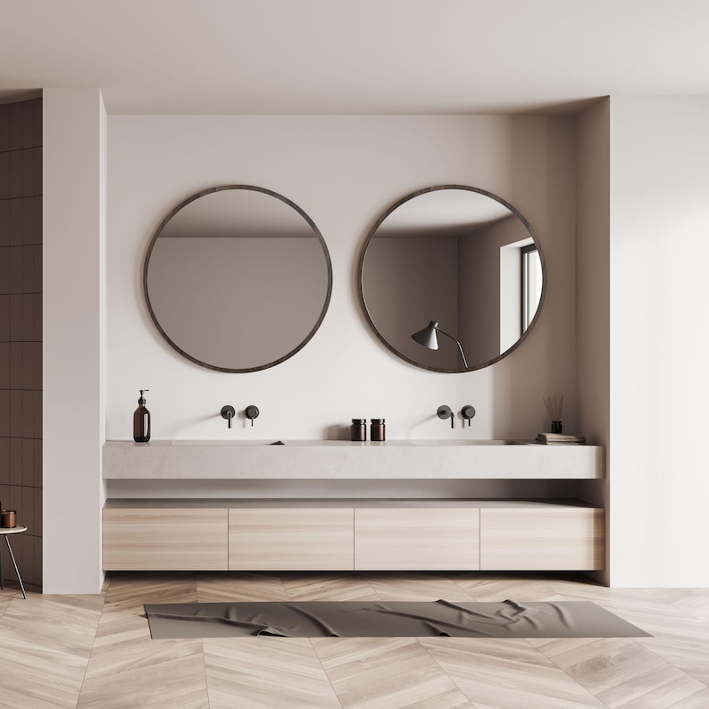 A modern bathroom design featuring two large, round mirrors with dark frames above a double vanity. The vanity has a light-colored countertop and wooden cabinets. Black faucets and a soap dispenser are visible.