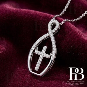 May include: A silver pendant necklace featuring a cross design. The pendant has an infinity symbol shape with a cross in the center, both encrusted with small, sparkling stones. The necklace is displayed on a dark red velvet surface.