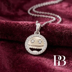 May include: A silver-toned circular pendant necklace featuring a stylized face design with two round-cut diamond eyes and a row of teeth. The outer edge of the circular frame is encrusted with small pavé-set diamonds, suspended from a delicate chain.