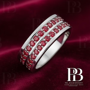 May include: A silver ring with two rows of small, vibrant red gemstones. The ring has a polished, reflective surface and is set against a deep red background. The design is elegant and eye-catching, showcasing the gemstones' rich color.