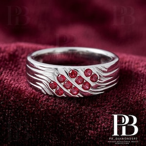 May include: A silver-toned band ring featuring a textured, wavy surface design. The ring is accented with eight small, round, vibrant red gemstones arranged in a diagonal pattern across the front of the band, creating a distinct visual contrast.