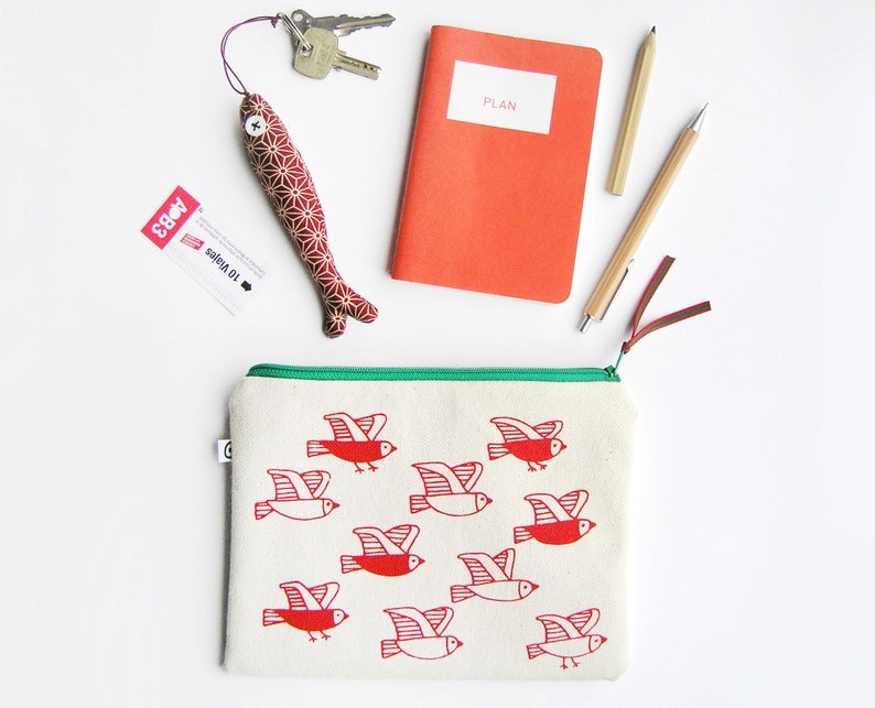 May include: A white canvas zippered pouch with a green zipper pull. The pouch is decorated with a red and white print of flying birds. The pouch is lying on a white surface.