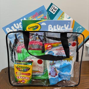 Puede incluir: Una bolsa transparente llena de materiales de arte, incluyendo crayones Crayola, libros de actividades de pegatinas y plastilina. La bolsa tiene asas negras y est&aacute; sobre una superficie de madera.