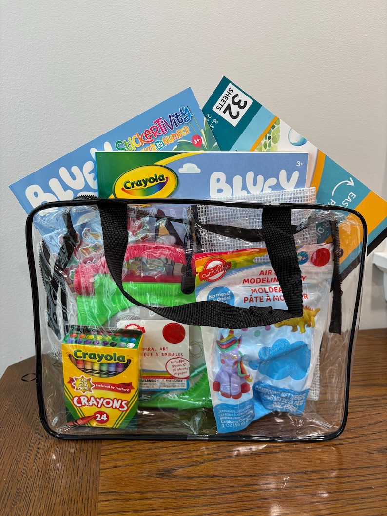 Puede incluir: Una bolsa transparente llena de materiales de arte, incluyendo crayones Crayola, libros de actividades de pegatinas y plastilina. La bolsa tiene asas negras y est&aacute; sobre una superficie de madera.