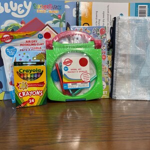 Puede incluir: Una colecci&oacute;n de materiales de arte para ni&ntilde;os, que incluye un kit de arte en espiral verde, crayones, arcilla de modelado que se seca al aire y libros de actividades con pegatinas. Los art&iacute;culos est&aacute;n colocados sobre una superficie de madera.