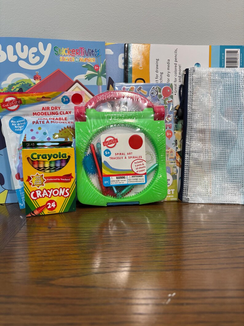 Puede incluir: Una colecci&oacute;n de materiales de arte para ni&ntilde;os, que incluye un kit de arte en espiral verde, crayones, arcilla de modelado que se seca al aire y libros de actividades con pegatinas. Los art&iacute;culos est&aacute;n colocados sobre una superficie de madera.