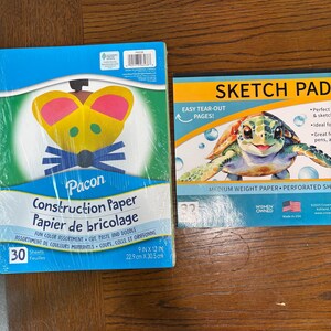 Puede incluir: Dos blocs de papel sobre una superficie de madera. Uno es un paquete azul de papel de construcci&oacute;n Pacon, 22,9 cm x 30,5 cm, con un dise&ntilde;o colorido. El otro es un bloc de dibujo con una ilustraci&oacute;n de tortuga y el texto "Sketch Pad".