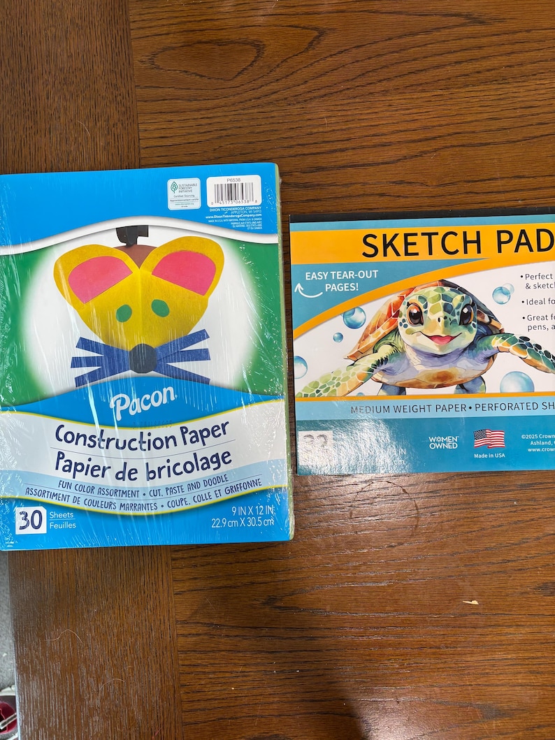 Puede incluir: Dos blocs de papel sobre una superficie de madera. Uno es un paquete azul de papel de construcci&oacute;n Pacon, 22,9 cm x 30,5 cm, con un dise&ntilde;o colorido. El otro es un bloc de dibujo con una ilustraci&oacute;n de tortuga y el texto "Sketch Pad".