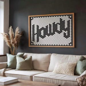May include: A living room with a beige sofa and green and cream cushions. A wooden-framed artwork with the word "Happy" in black and white hexagon tiles hangs on the dark grey wall. Pampas grass and vases are on a nearby windowsill.
