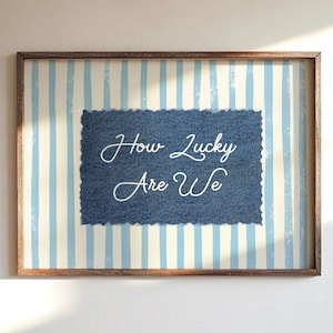 May include: Framed wall art with a blue and white striped background. A denim-textured square in the center features the cursive text "How Lucky Are We" in white. The frame is a dark brown color.