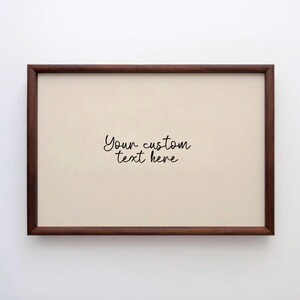 May include: A rectangular framed print with a dark brown wooden frame. The print has a cream-coloured background with the handwritten text "Your custom text here" in black cursive font.