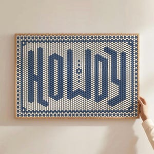 May include: Framed wall art featuring the word "HOWDY" in a blue mosaic design. The background is a mix of white and light blue mosaic tiles. The frame is a light brown colour.
