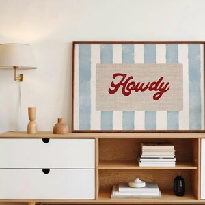 May include: A framed print featuring the word "Howdy" in red script on a beige rectangle, set against a backdrop of blue and white vertical stripes. The artwork is displayed above a wooden cabinet with white drawers and open shelves.