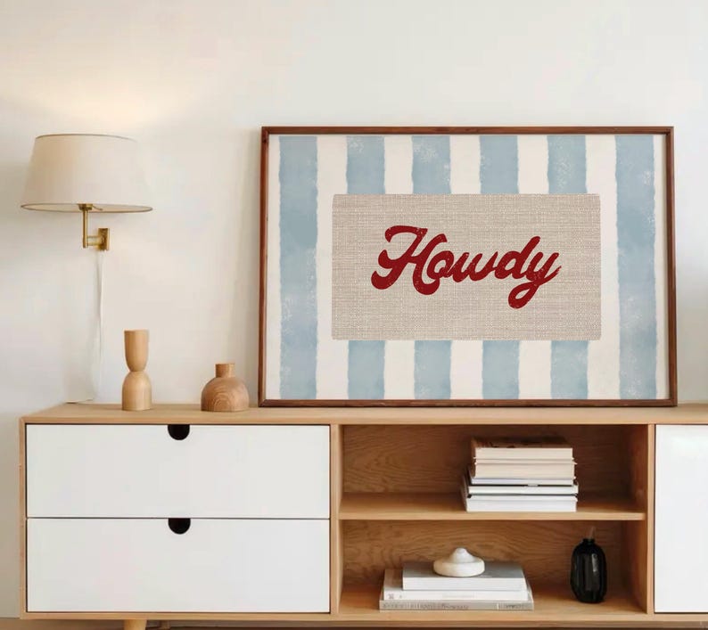 May include: A framed print featuring the word "Howdy" in red script on a beige rectangle, set against a backdrop of blue and white vertical stripes. The artwork is displayed above a wooden cabinet with white drawers and open shelves.