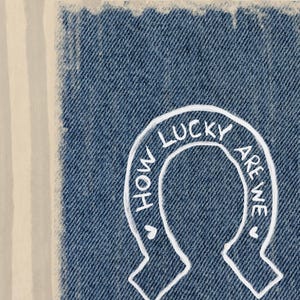 May include: Close-up of a denim patch with a white horseshoe design. The text "HOW LUCKY ARE WE" is written inside the horseshoe. The denim has a frayed edge and is set against a striped background.