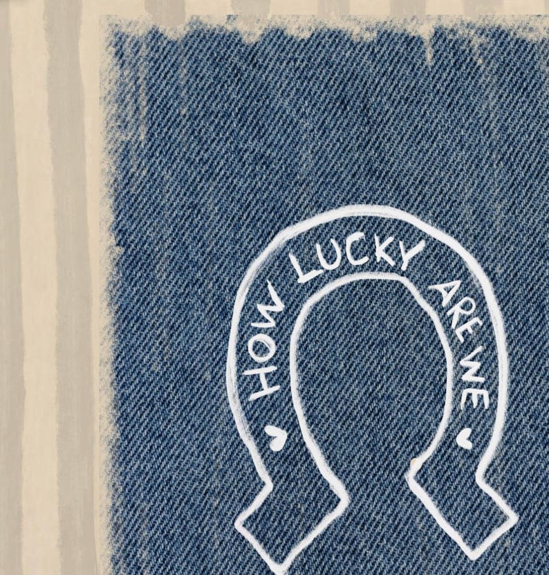 May include: Close-up of a denim patch with a white horseshoe design. The text "HOW LUCKY ARE WE" is written inside the horseshoe. The denim has a frayed edge and is set against a striped background.