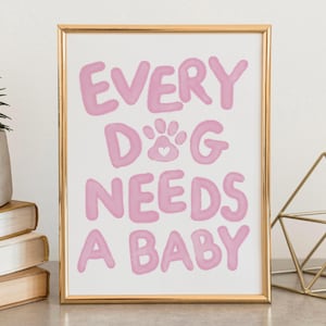 May include: A framed print with the text "EVERY DOG NEEDS A BABY" in pink bubble letters. The print includes a paw print with a heart in the center. The frame is gold, and the print is displayed on a gray surface.