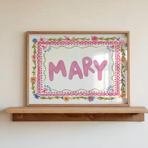 Może przedstawiać: Oprawiona grafika z imieniem "MARY" w r&oacute;żowych literach. Grafika otoczona jest kwiatową obw&oacute;dką z r&oacute;żowymi, zielonymi, niebieskimi i ż&oacute;łtymi akcentami. Rama jest w jasnobrązowym kolorze, a grafika jest eksponowana na drewnianej p&oacute;łce.