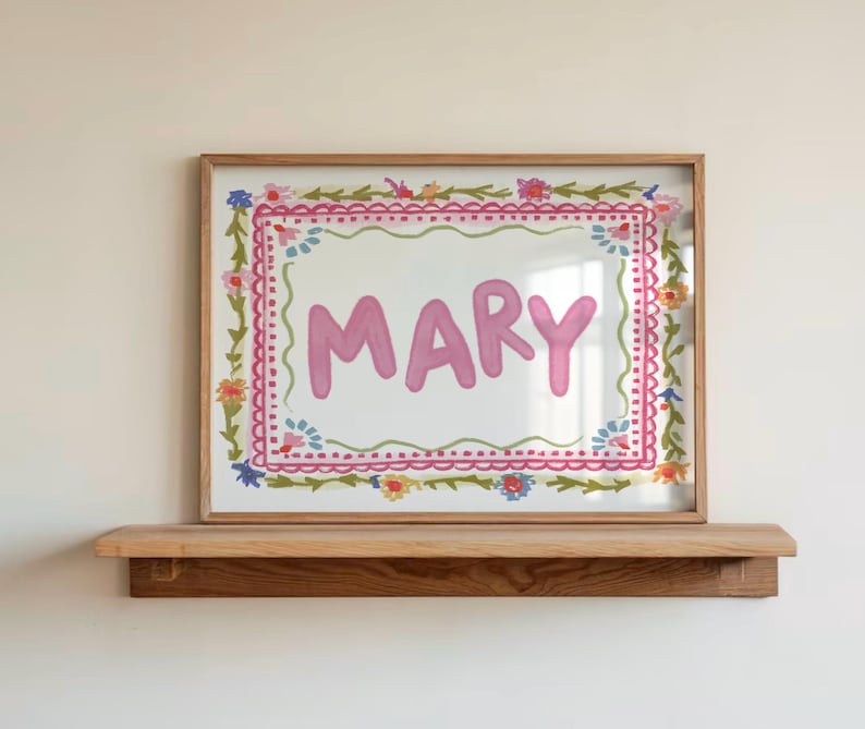 Może przedstawiać: Oprawiona grafika z imieniem "MARY" w r&oacute;żowych literach. Grafika otoczona jest kwiatową obw&oacute;dką z r&oacute;żowymi, zielonymi, niebieskimi i ż&oacute;łtymi akcentami. Rama jest w jasnobrązowym kolorze, a grafika jest eksponowana na drewnianej p&oacute;łce.