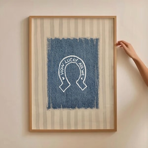May include: Framed artwork with a horseshoe design and the text "HOW LUCKY ARE WE" in white on a blue denim-style background. The frame is light brown, and the background has vertical stripes.