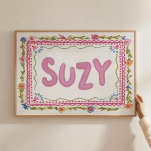Może przedstawiać: Oprawione dzieło sztuki z imieniem "SUZY" w kolorze r&oacute;żowym, otoczone kwiatową obw&oacute;dką. Grafika ma białe tło z r&oacute;żowo-zielonym kwiatowym wzorem. Rama wykonana jest z jasnobrązowego drewna.