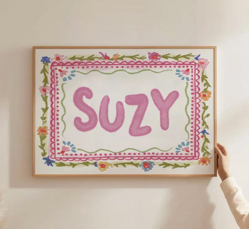 Może przedstawiać: Oprawione dzieło sztuki z imieniem "SUZY" w kolorze r&oacute;żowym, otoczone kwiatową obw&oacute;dką. Grafika ma białe tło z r&oacute;żowo-zielonym kwiatowym wzorem. Rama wykonana jest z jasnobrązowego drewna.