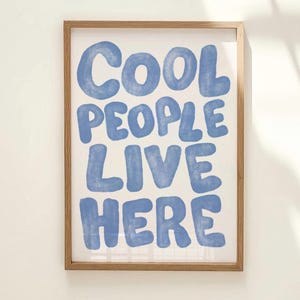 Può includere: Una stampa artistica incorniciata con il testo "COOL PEOPLE LIVE HERE" in grandi lettere arrotondate blu chiaro. La stampa è su sfondo bianco e incorniciata in una cornice di legno naturale, adatta all'arredamento della casa.