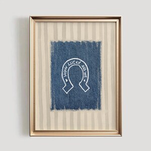May include: Framed artwork featuring a blue denim square with a white horseshoe design. The text "HOW LUCKY ARE WE" is written inside the horseshoe. The frame is gold, and the background has vertical beige and white stripes.