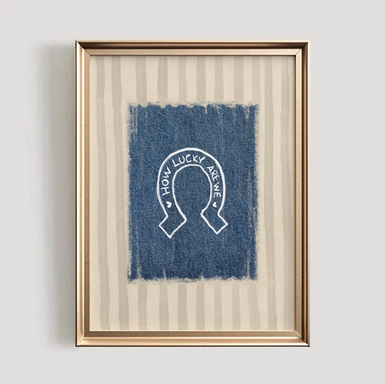 May include: Framed artwork featuring a blue denim square with a white horseshoe design. The text "HOW LUCKY ARE WE" is written inside the horseshoe. The frame is gold, and the background has vertical beige and white stripes.