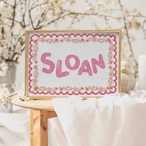 Może przedstawiać: Oprawiony wydruk z imieniem "SLOAN" w r&oacute;żowych literach. Wydruk ma falisty r&oacute;żowy obramowanie i wz&oacute;r girlandy kwiatowej. Rama jest złota.