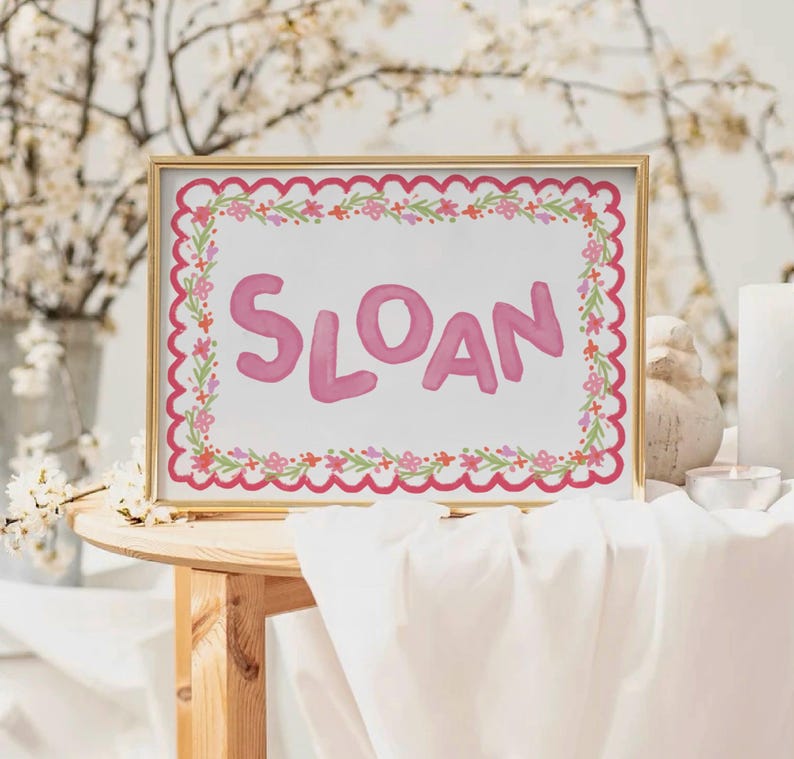 Może przedstawiać: Oprawiony wydruk z imieniem "SLOAN" w r&oacute;żowych literach. Wydruk ma falisty r&oacute;żowy obramowanie i wz&oacute;r girlandy kwiatowej. Rama jest złota.