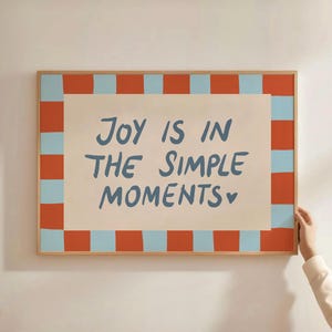 以下が含まれることがあります： 青い文字で「JOY IS IN THE SIMPLE MOMENTS」と書かれた額入りのアートプリント。 アートワークは、クリーム色の背景に、オレンジと水色の正方形が交互に並んだボーダーがあります。 フレームは明るい茶色です。