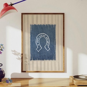 May include: Framed artwork featuring a blue denim-textured square with a white horseshoe design and the text "HOW LUCKY ARE WE" in a circular arrangement. The artwork is set against a striped background and framed in a wooden frame.